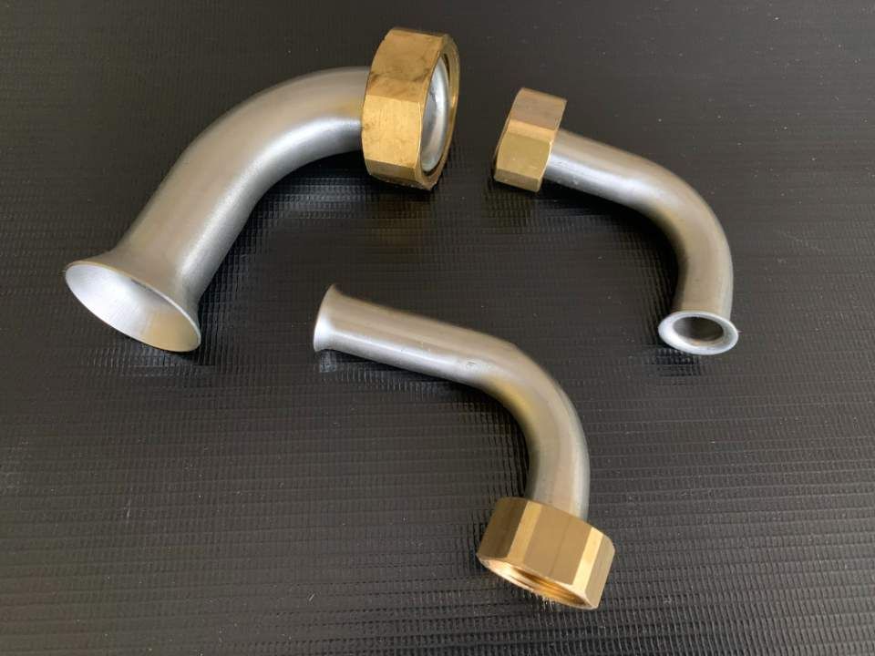 Drei gebogene, silberne Metallrohrverbinder mit Messingsechskantenden auf dunklem Hintergrund