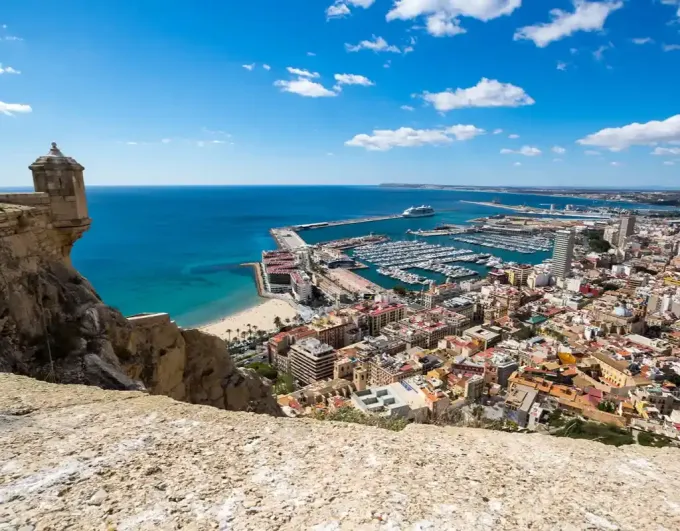 Playa soleada con sombrillas y tumbonas con vistas a un pueblo portuario y una fortaleza en la cima de una colina.