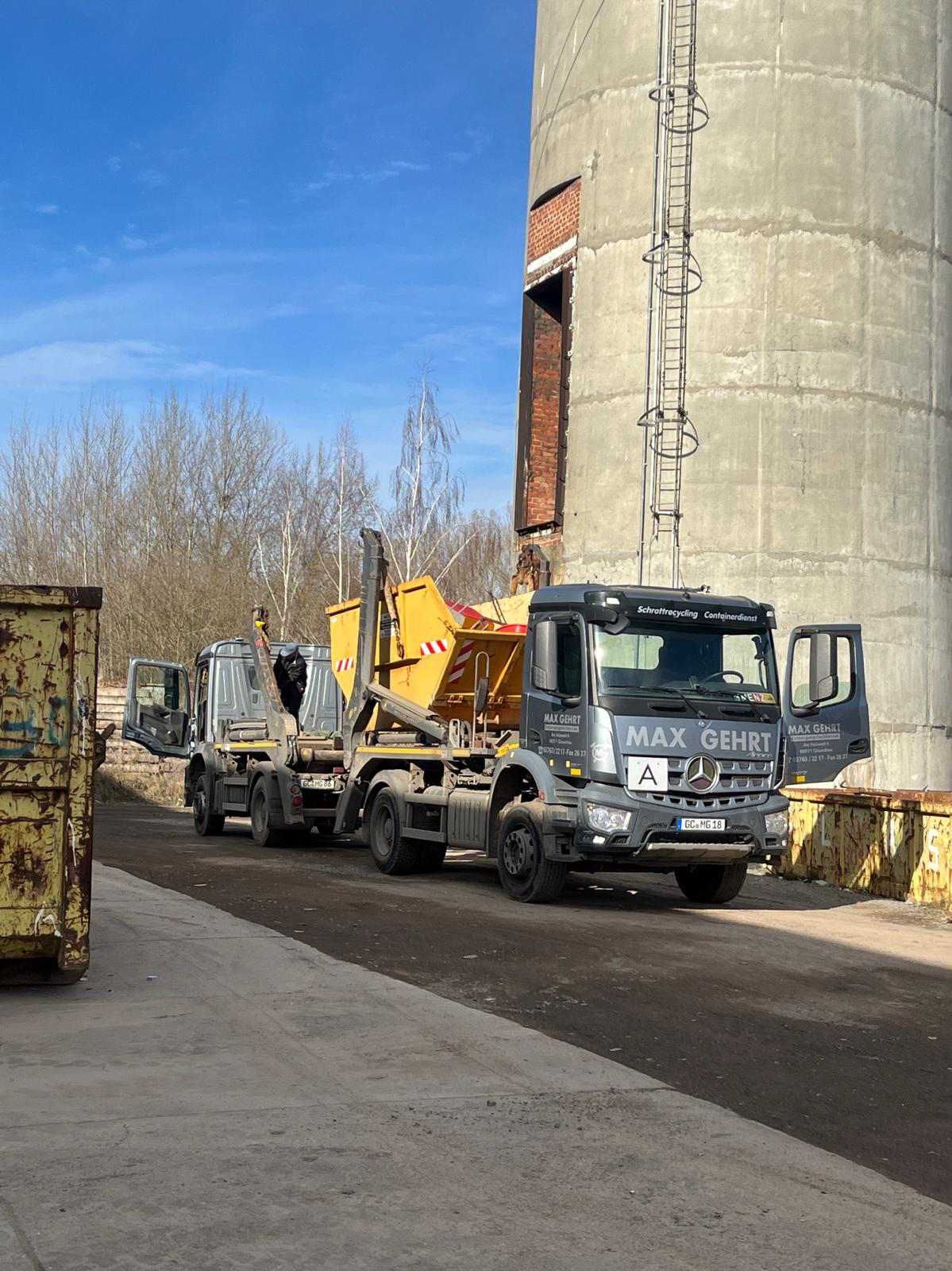 Ein Lastwagen transportiert einen gelben Bagger neben einem hohen Betonsilo auf einer Industriestraße.