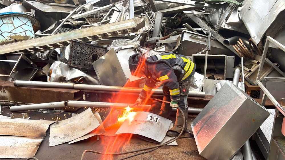 Feuerwehrmann bekämpft mit einem Schlauch Flammen inmitten eines Haufens Metalltrümmer und Wrackteilen.