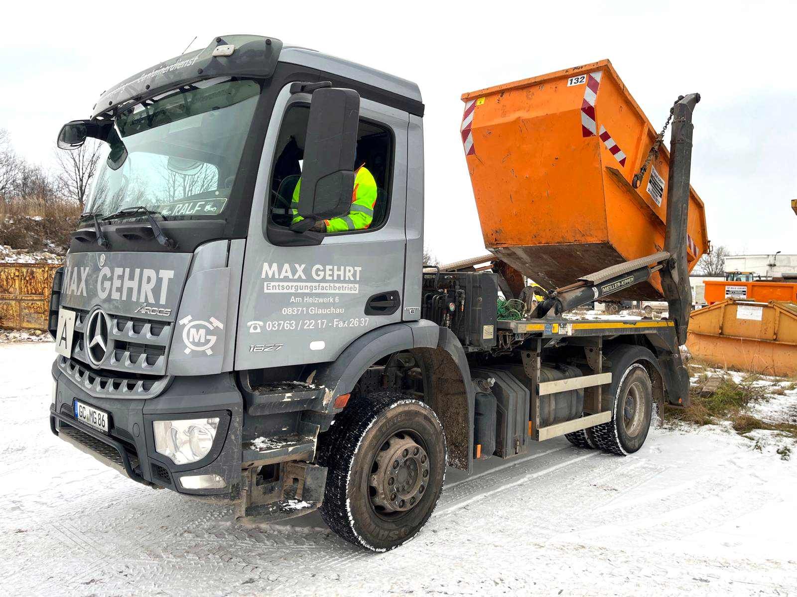 Ein grauer Mercedes-Benz-Lkw mit einem orangefarbenen Abrollcontainer auf der Ladefläche steht auf schneebedecktem Boden.