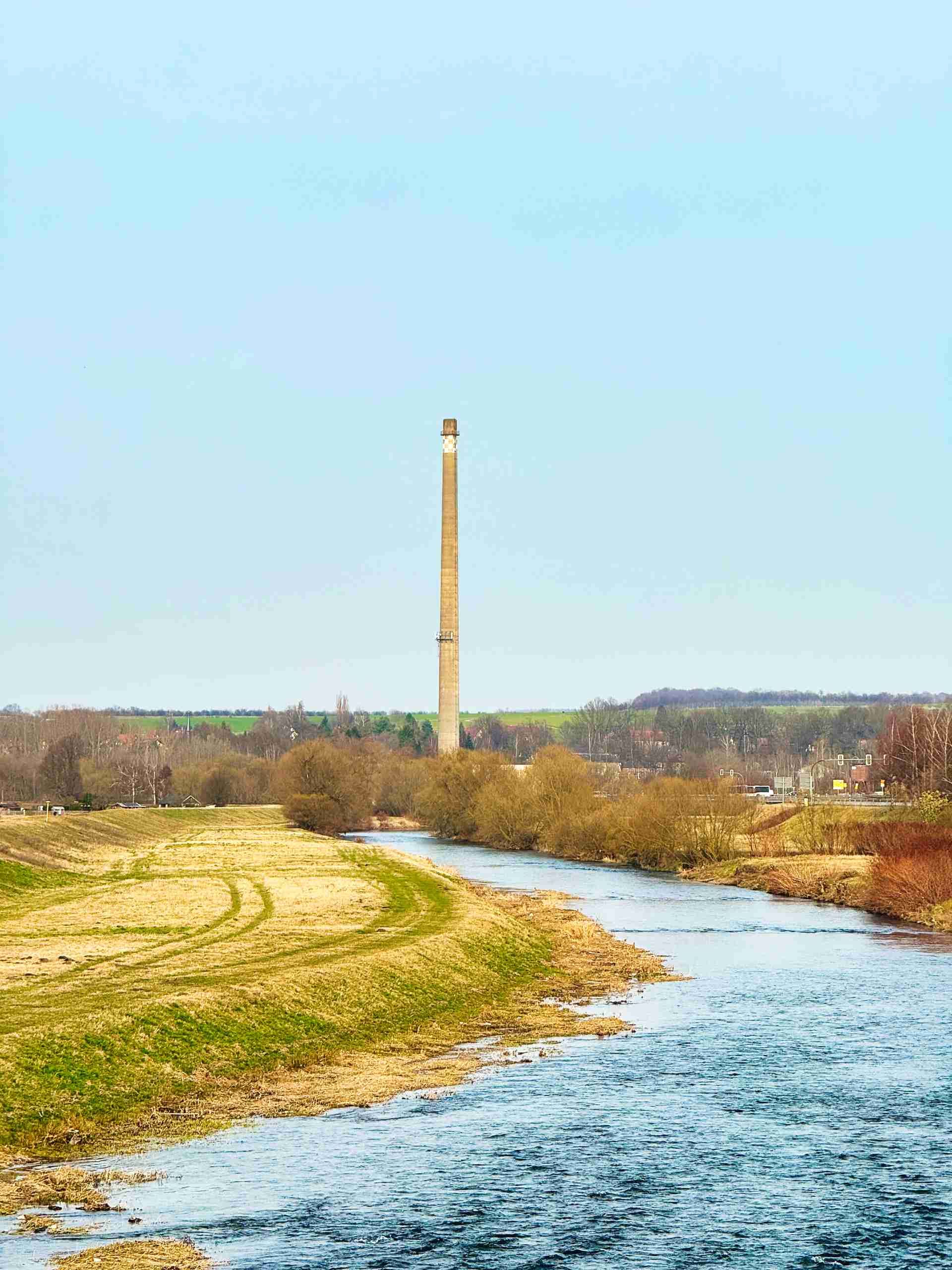 Ein Fluss schlängelt sich durch eine ländliche Landschaft, unter dem sich ein hoher Steinkamin erhebt, unter einem klaren blauen Himmel