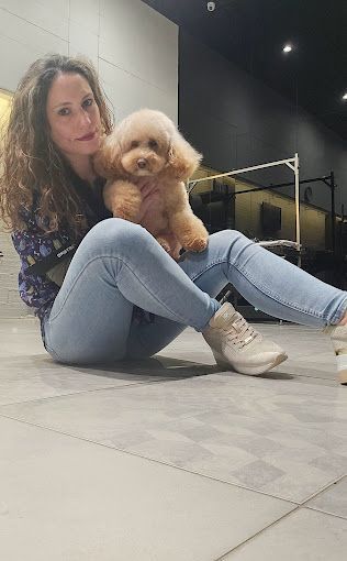Una mujer con vaqueros está sentada en el suelo, sosteniendo un perrito marrón. Sonríe, en el interior.
