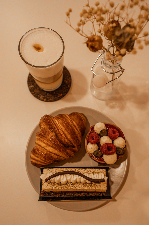 Café et viennoiseries sur une table avec un croissant, une tarte, un gâteau, un café latte et des fleurs séchées dans un vase.