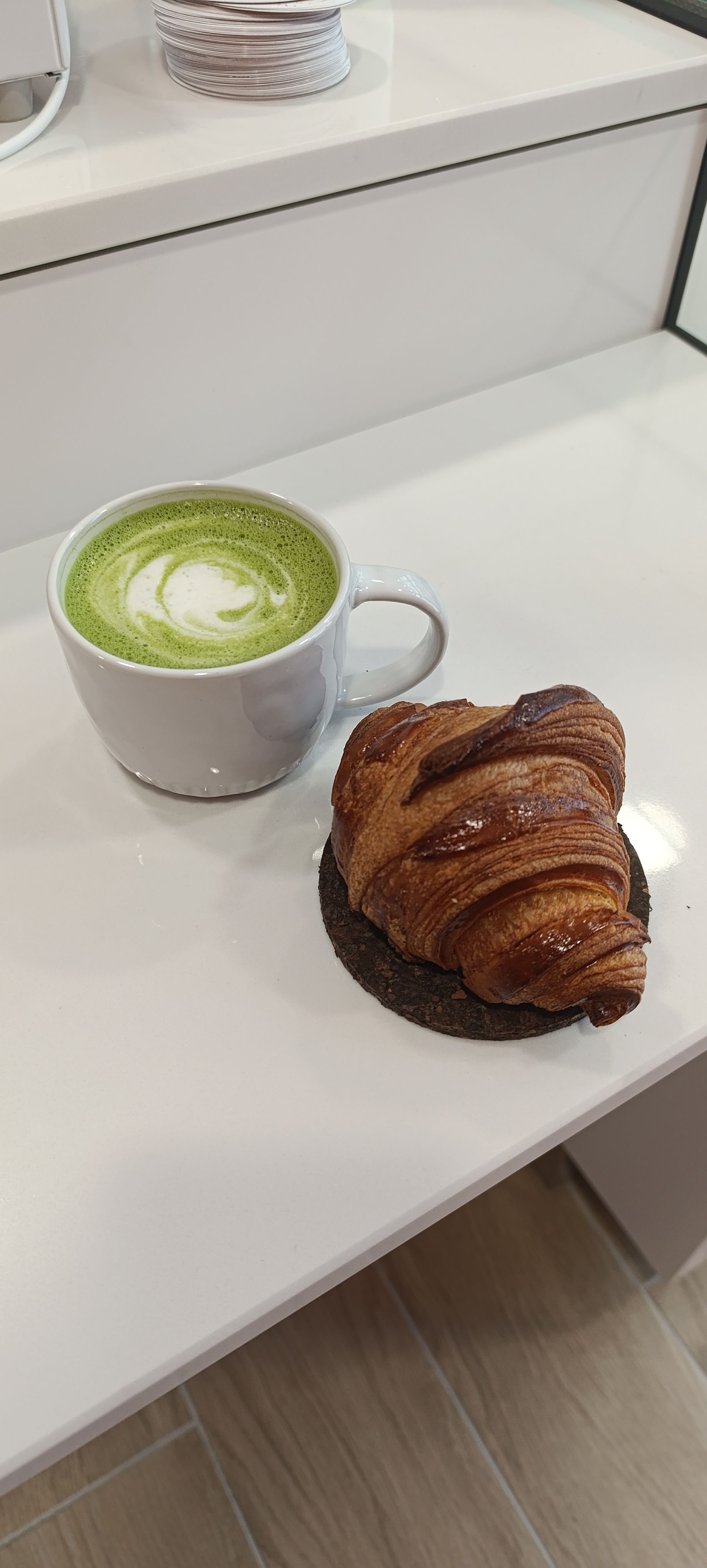 Une tasse de matcha latte vert avec un croissant sur une table blanche près d'une fenêtre.