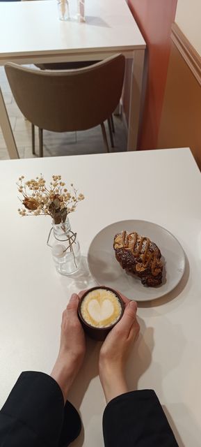 Des mains tenant un café et une viennoiserie à une table de café blanche, avec un petit vase de fleurs séchées.