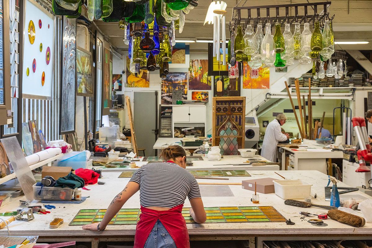 Femme travaillant sur un projet en verre dans un atelier coloré ; diverses pièces de verre, outils et œuvres d’art.