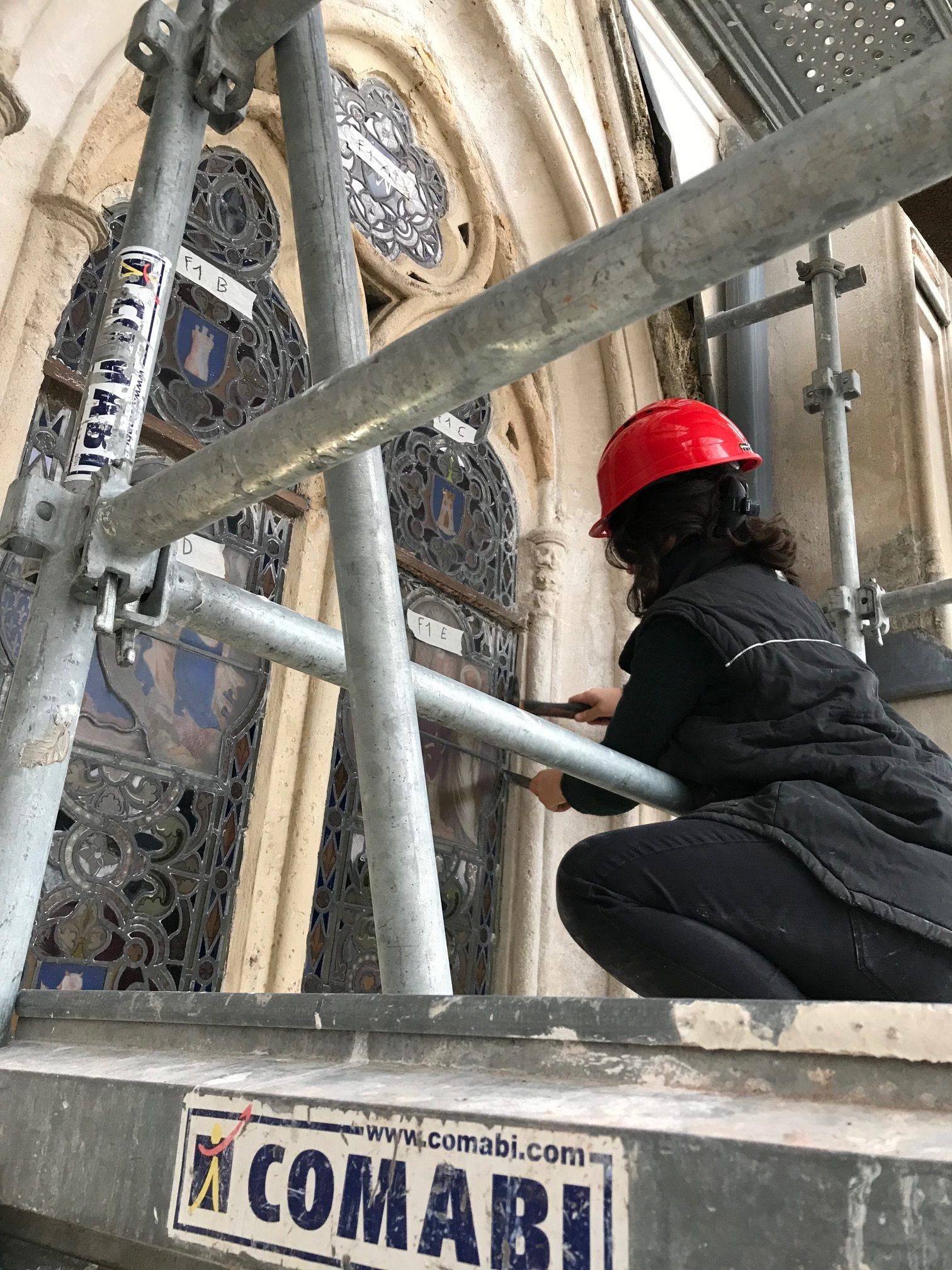 Une personne portant un casque rouge sur un échafaudage inspecte un vitrail.