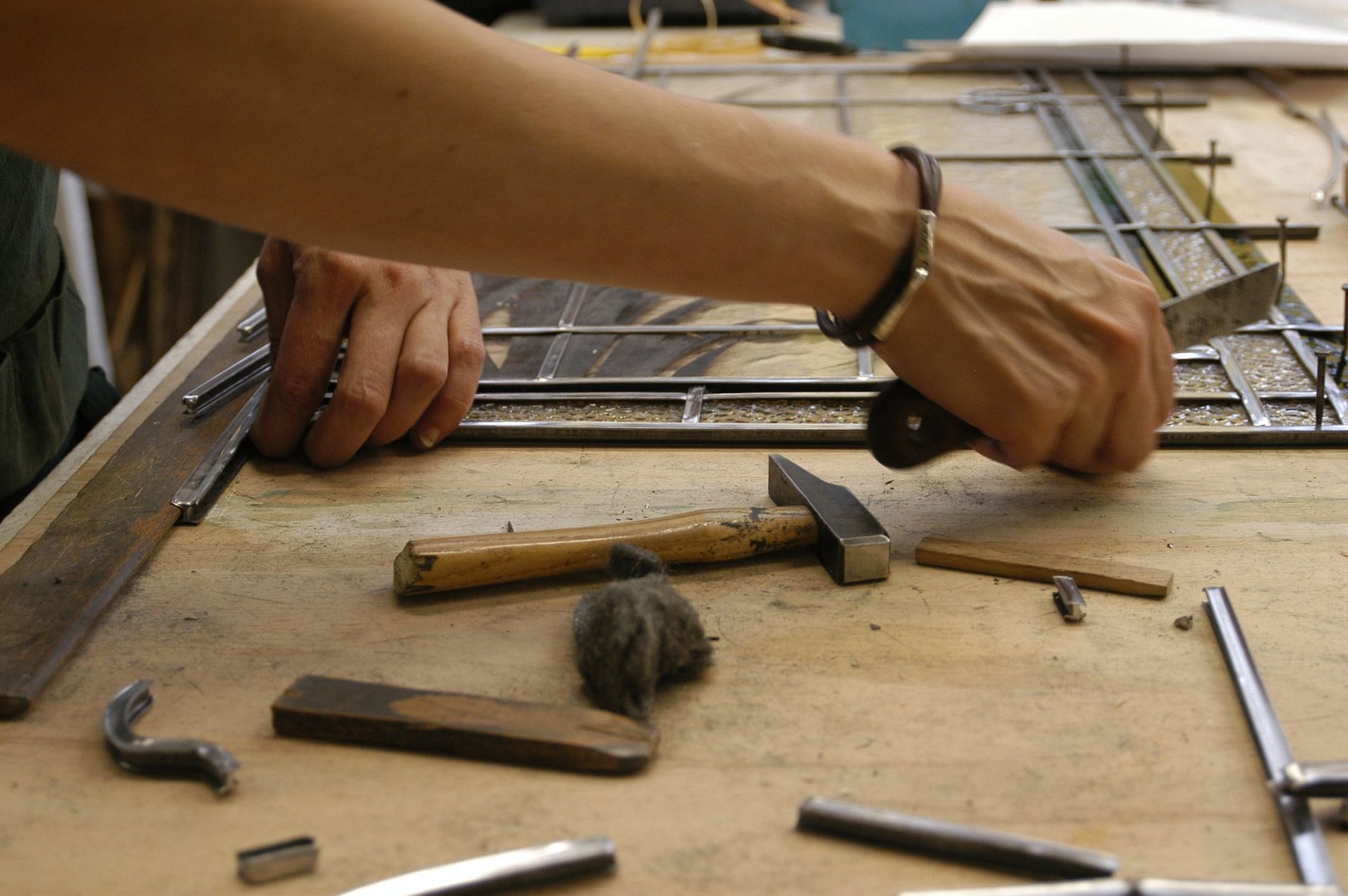 Personne travaillant à la réalisation d'un vitrail, mains visibles. Marteau, pièces métalliques et outils sur une surface en bois.