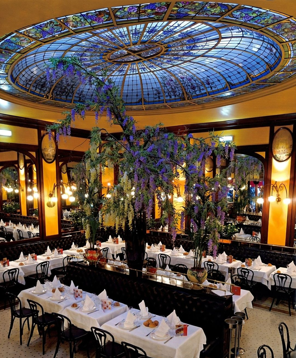 Intérieur de restaurant élégant avec des tables nappées de blanc, un plafond en dôme de vitraux et une glycine.