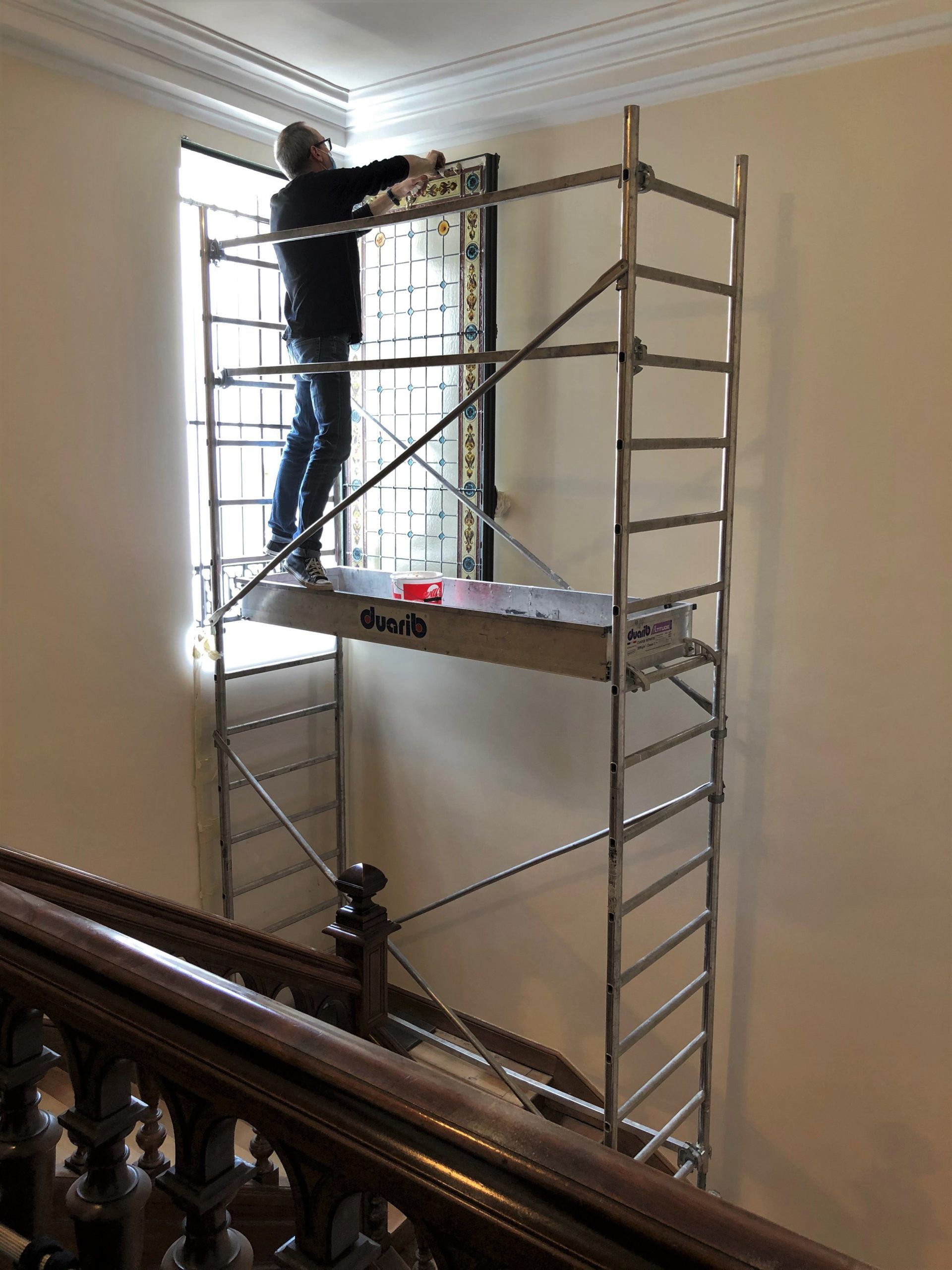Une personne se tient sur un échafaudage, travaillant sur une fenêtre à barreaux. À l'intérieur, près d'un escalier.