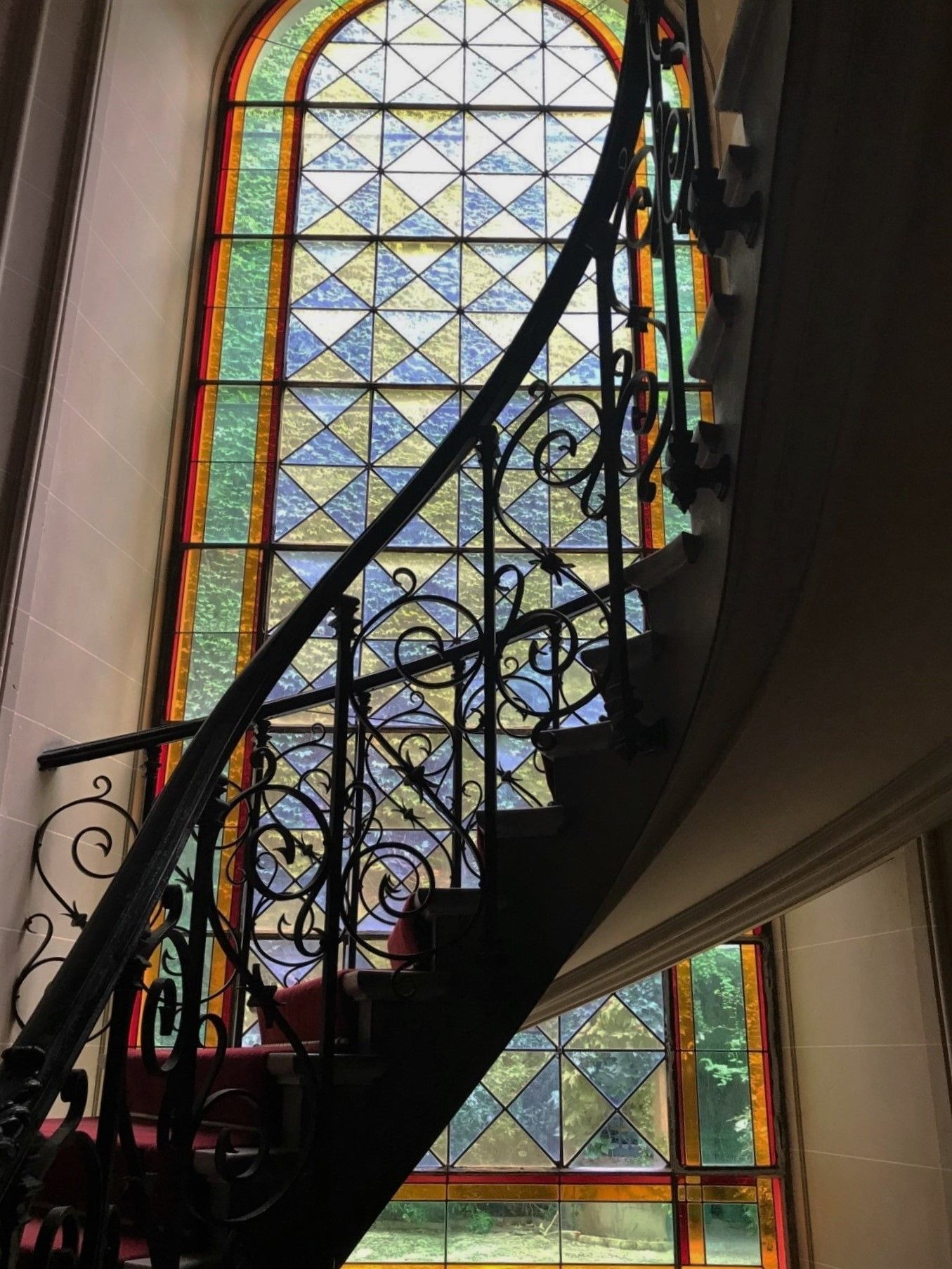 Escalier en colimaçon avec une rampe noire ornée, près d'un grand vitrail aux motifs géométriques bleus et jaunes.