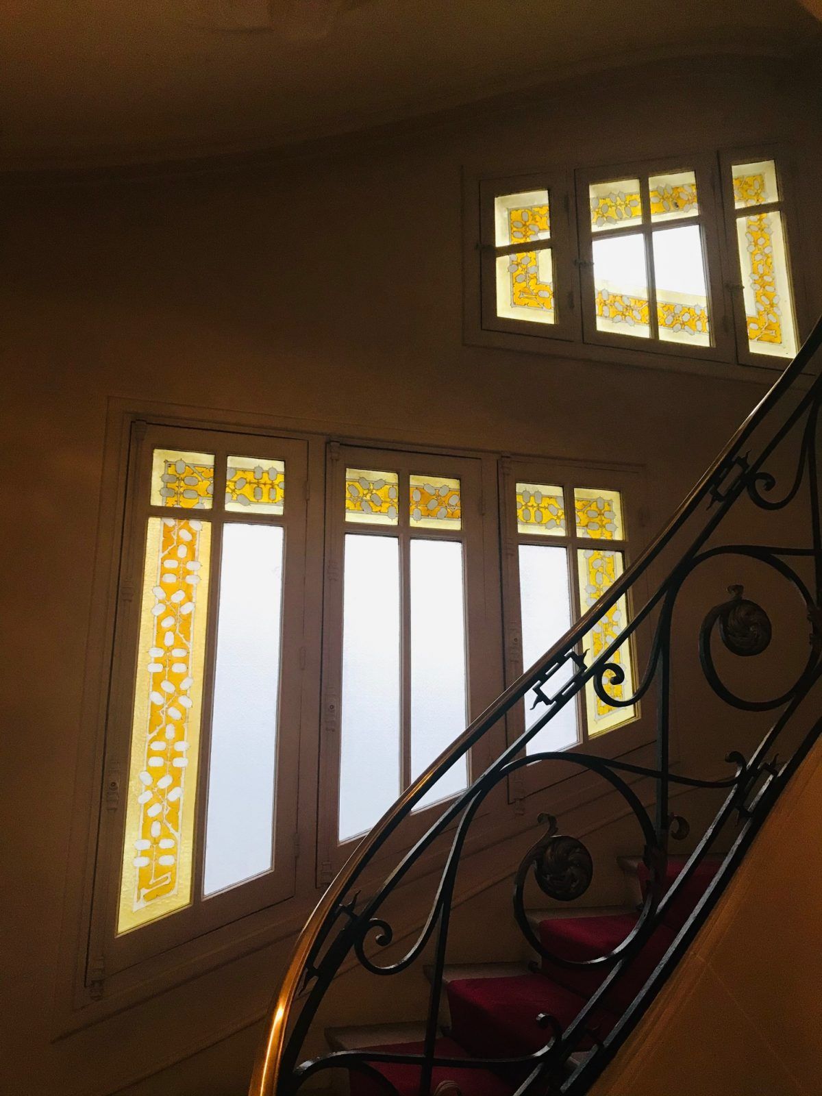 Escalier avec rampe en fer forgé, lumière du soleil filtrant à travers les vitraux, moquette rouge.