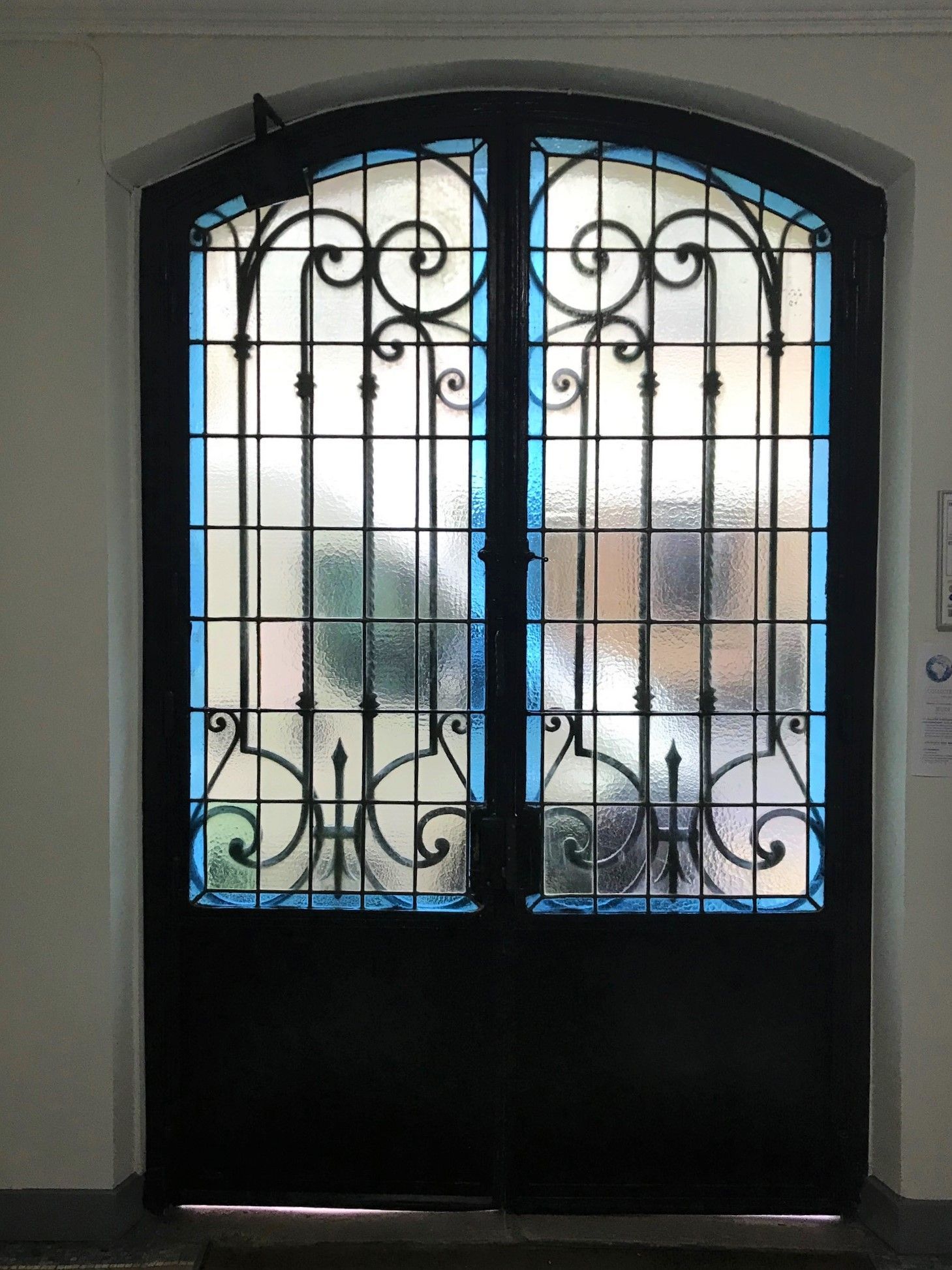 Double porte en métal noir avec grilles ornementées et vitres dépolies, garniture bleue, encastrée dans un mur blanc.
