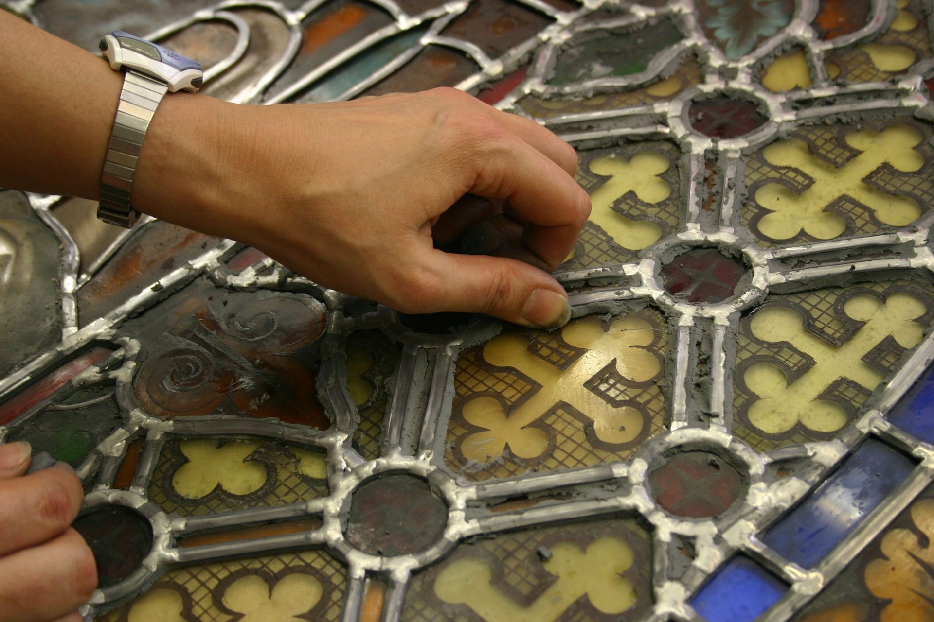 Main travaillant sur un vitrail, en retirant un petit morceau. Montre en argent au poignet.