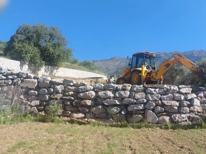 Excavadora amarilla detrás de un muro de piedra en un campo de hierba bajo un cielo azul.