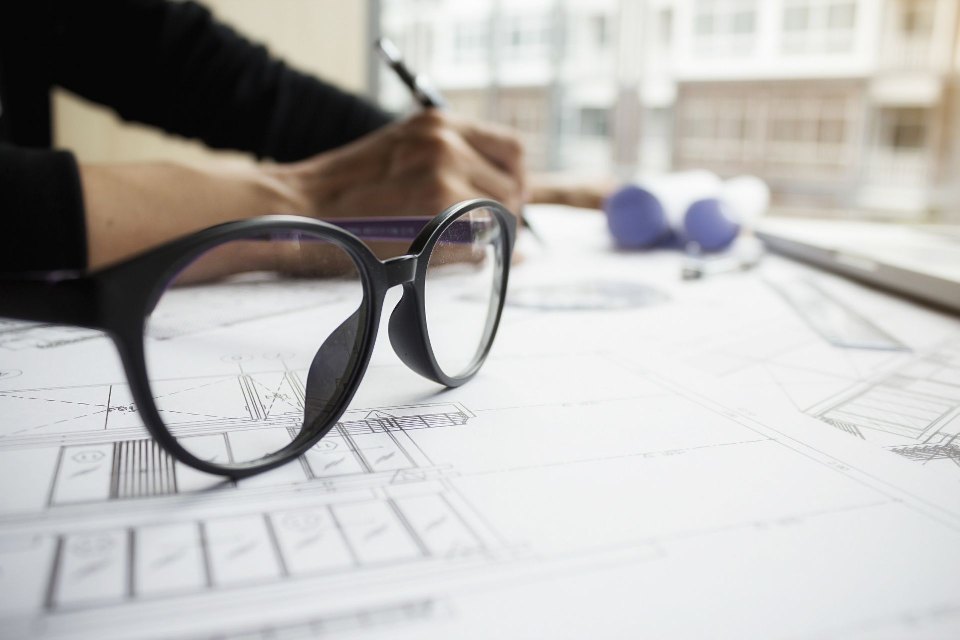 Pessoa assinando um documento em uma mesa; outras duas pessoas aparecem desfocadas ao fundo.
