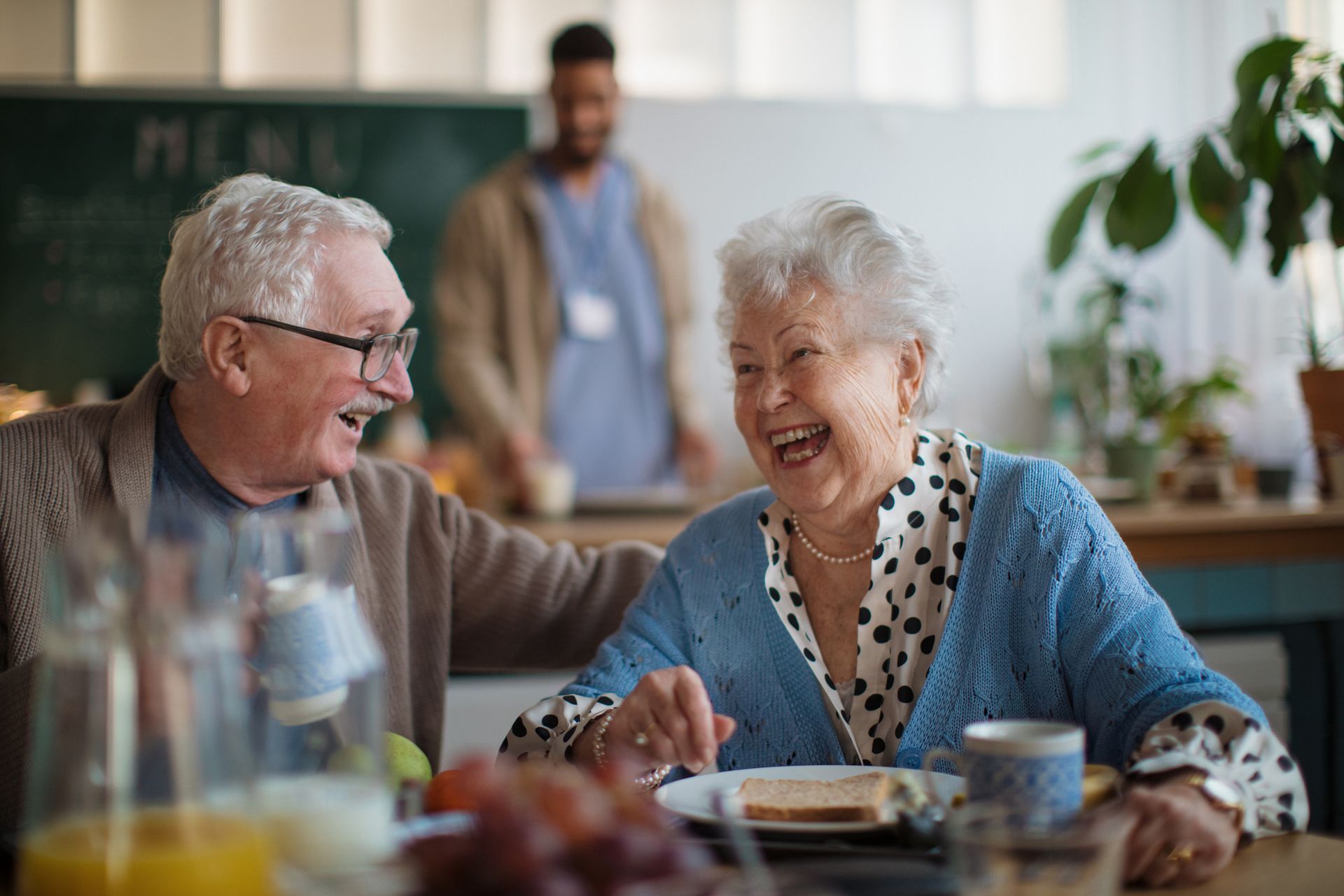 Pareja de ancianos riendo, comiendo en una mesa, con un cuidador al fondo.