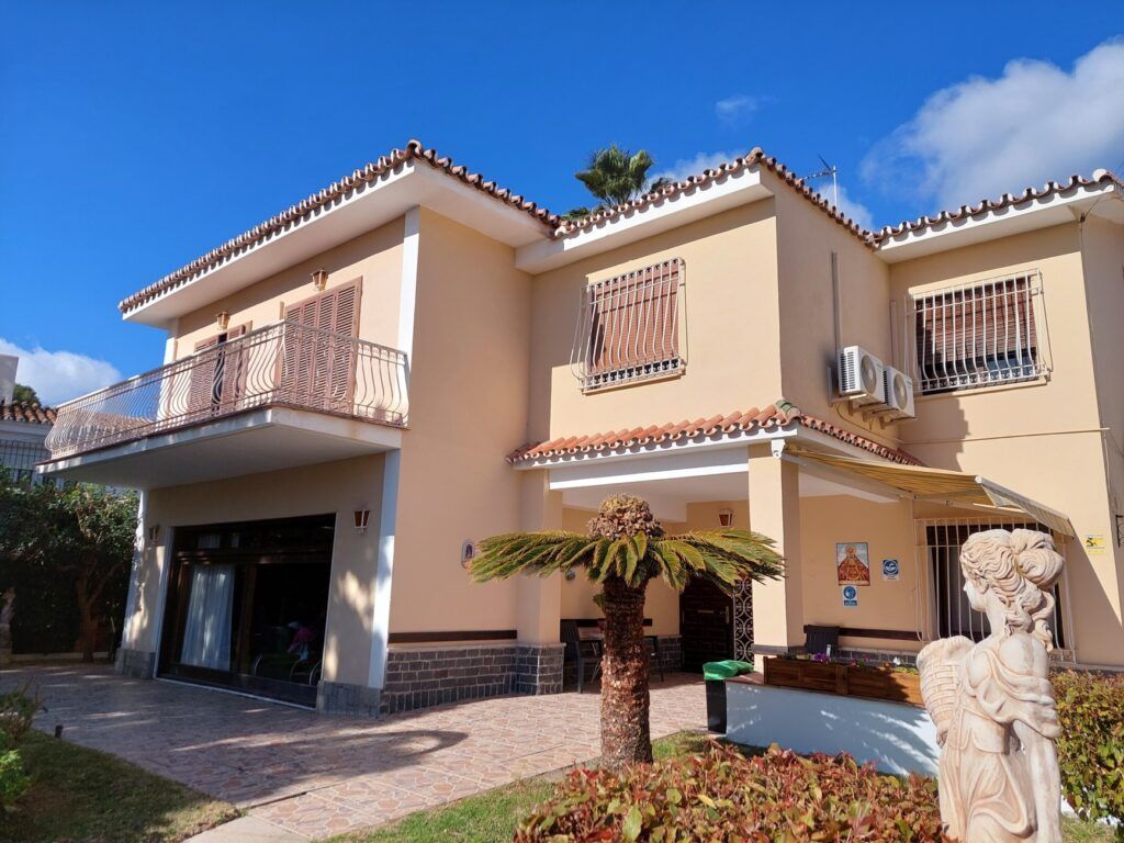 Casa beige de dos pisos con techo de tejas marrones y balcón bajo un cielo azul.