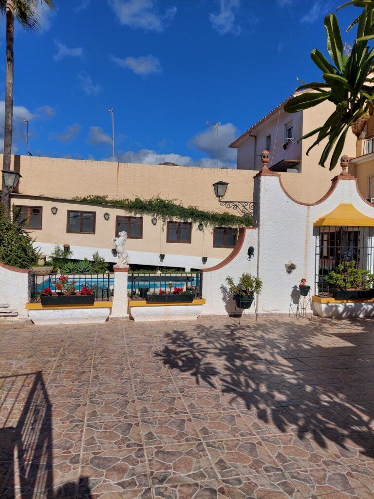Una plaza soleada con edificios, plantas y una estatua, con un cielo azul encima.