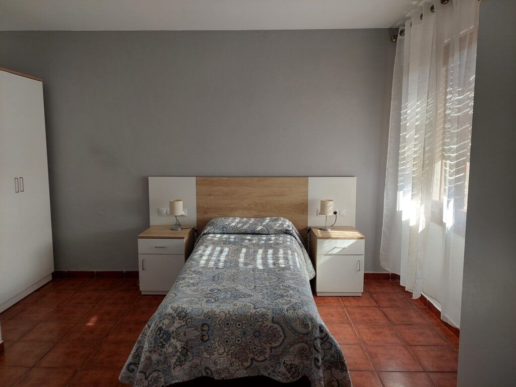 Dormitorio con cama individual, mesitas de noche y armario. Paredes grises, suelo marrón y una ventana con cortinas blancas.
