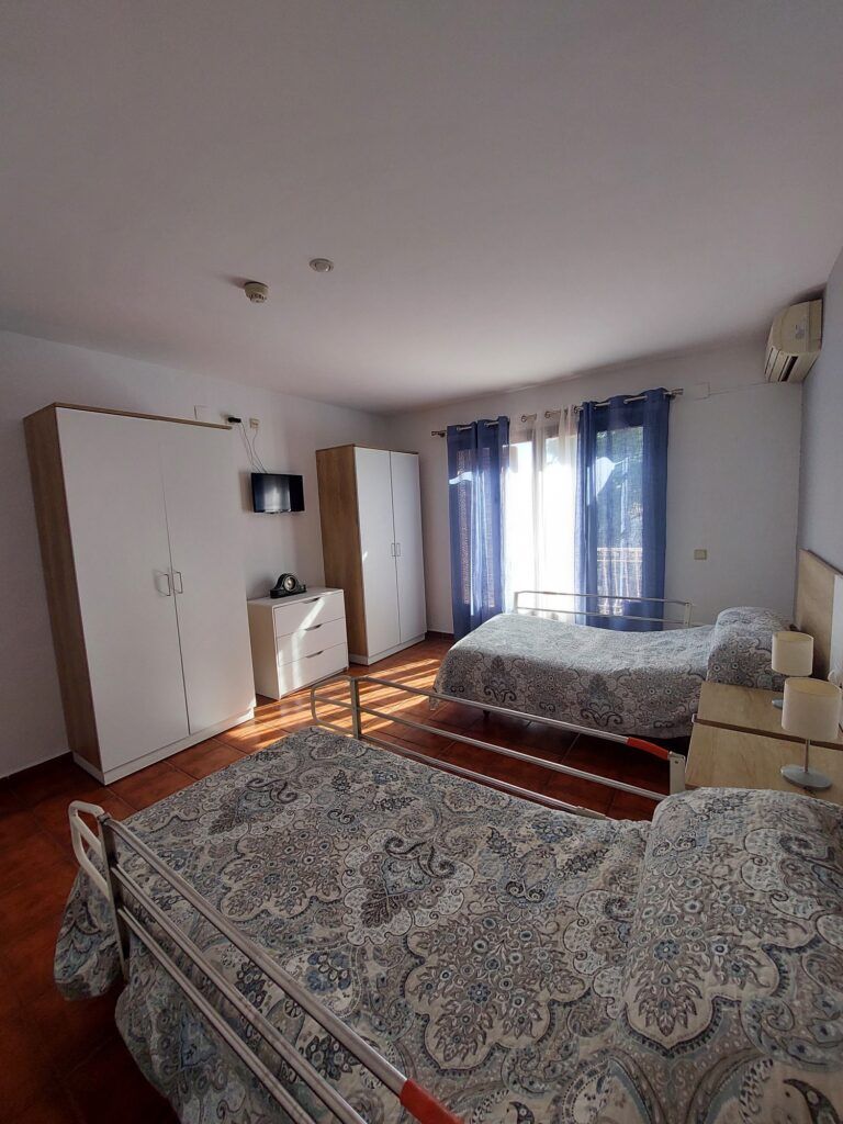 Dormitorio con dos camas individuales, dos armarios, cómoda y una ventana con cortinas azules; colores neutros.