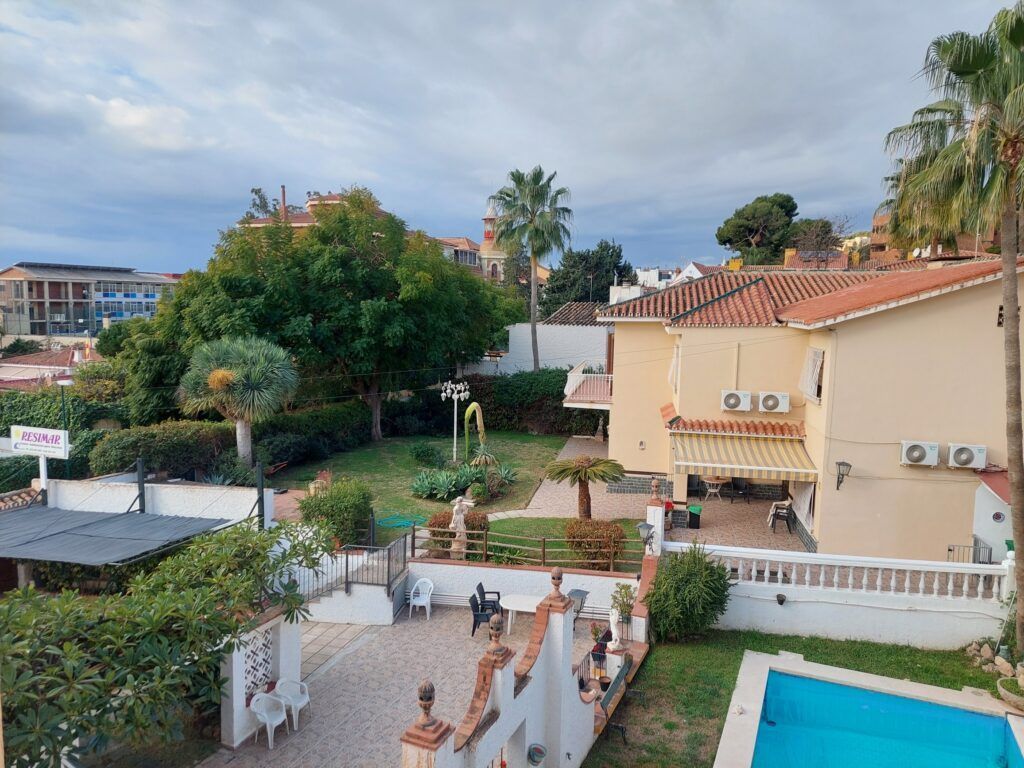 Vista de una casa con tejado de tejas rojas, jardín, piscina y palmeras. Cielo nublado al fondo.