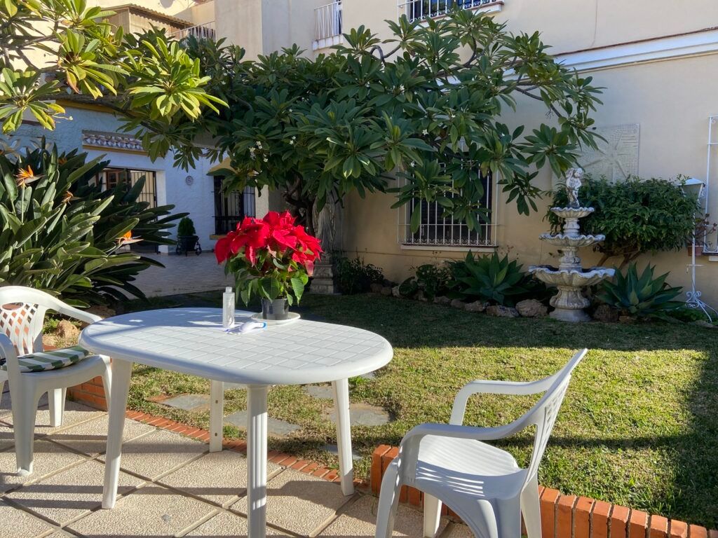 Mesa y sillas blancas en un patio, flor de Pascua roja, fuente y jardín en un entorno soleado.