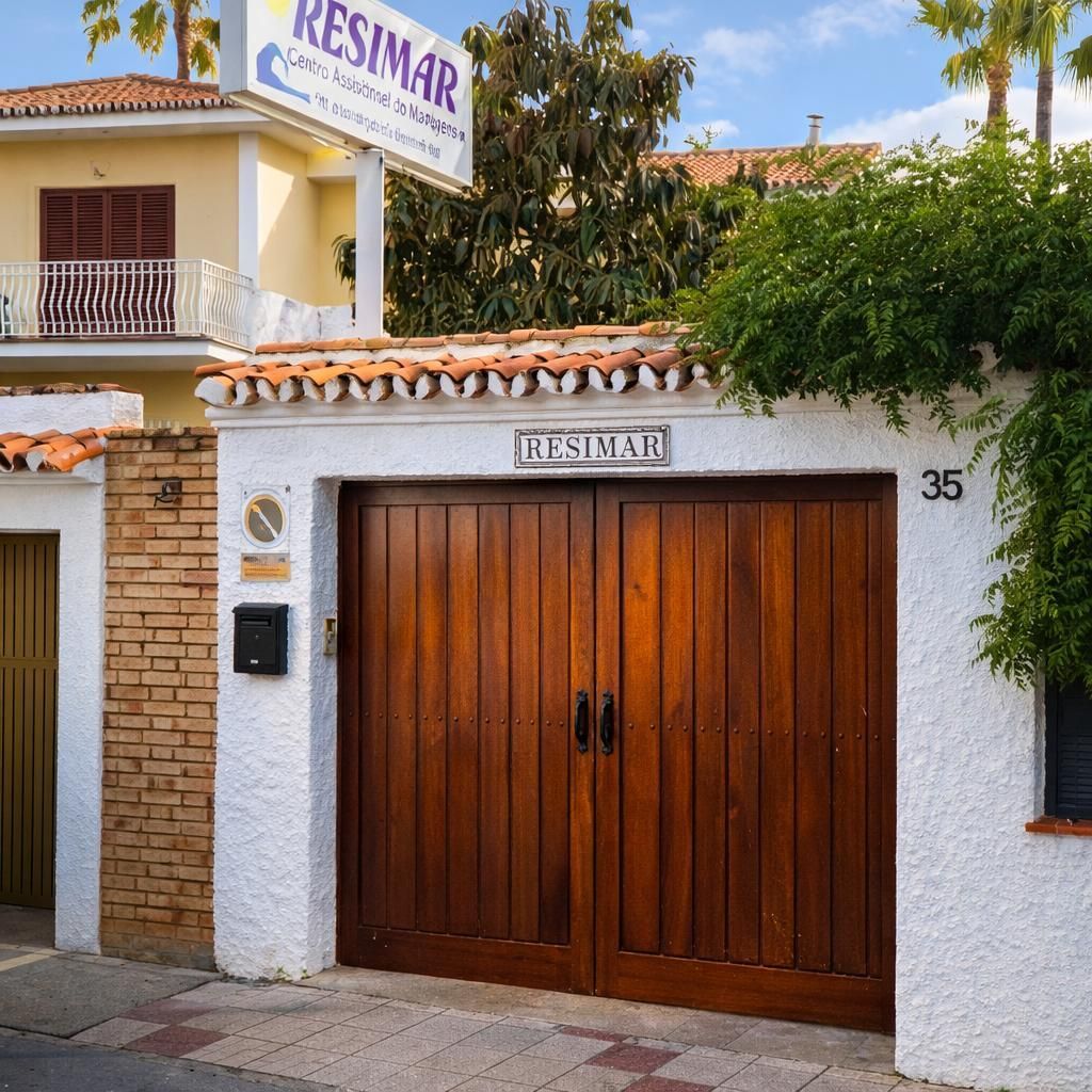 Un letrero comercial de RESIMAR cuelga sobre una pared blanca con una gran puerta de madera en el número 35, a la sombra de la vegetación.