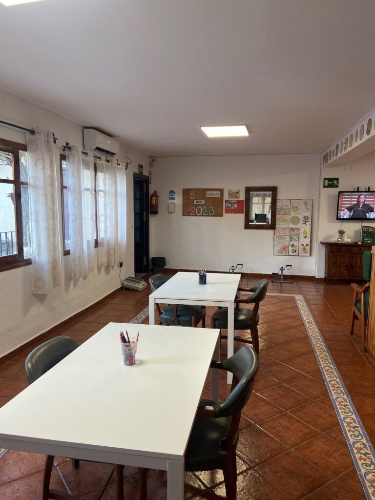 Un aula con mesas blancas y sillas negras, ventanas con cortinas blancas y un televisor en la pared.