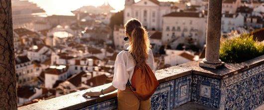 Une femme avec un sac à dos contemple une ville ensoleillée depuis une terrasse carrelée au coucher du soleil.