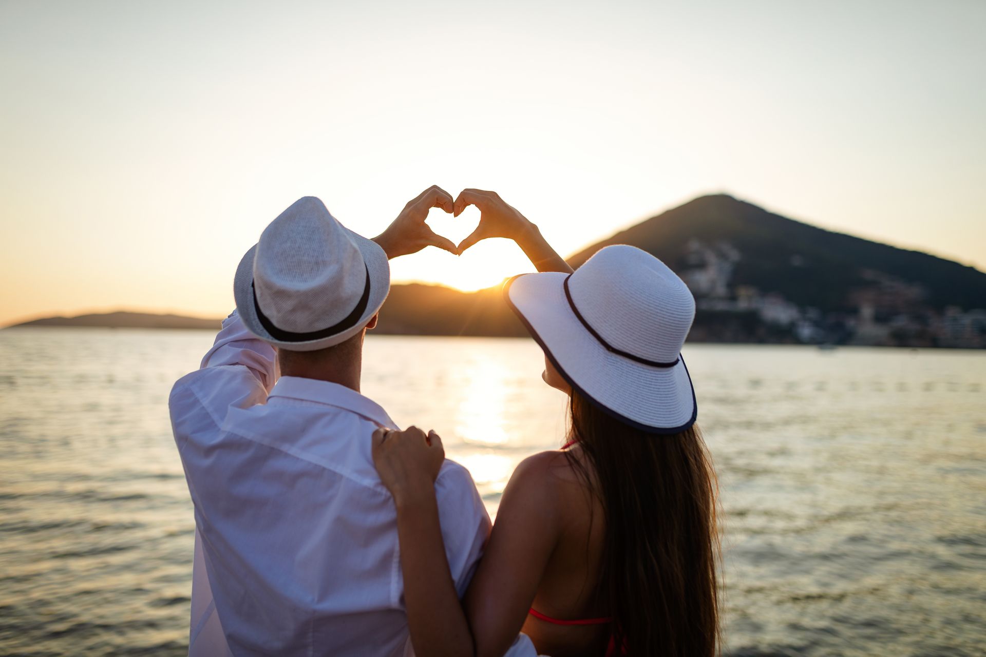 Deux personnes portant des chapeaux blancs forment un cœur avec leurs mains au coucher du soleil, au bord de l’eau.