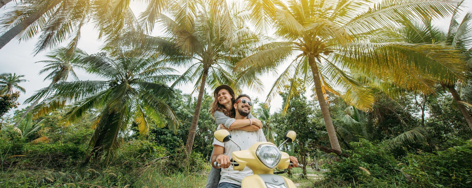 Un couple posant sur un scooter blanc dans un jardin tropical luxuriant, sous des palmiers.