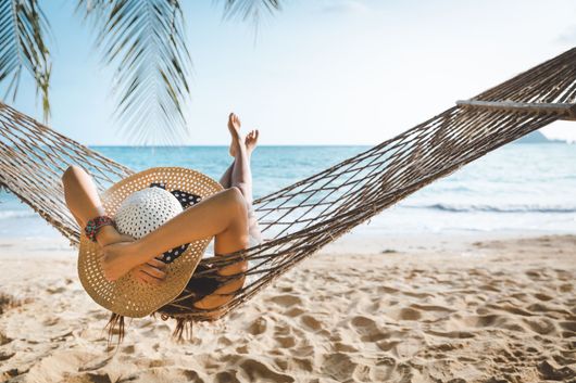 Personne se détendant dans un hamac sur une plage ensoleillée, les pieds en l’air, avec l’océan en arrière-plan.
