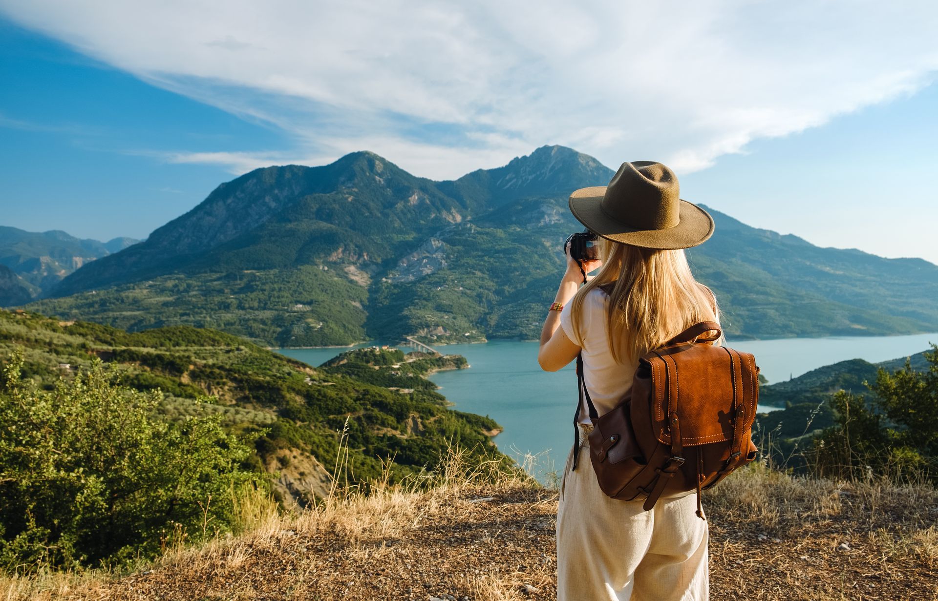 Une personne avec un sac à dos et un chapeau contemple un paysage de montagnes verdoyantes et un lac sous un ciel bleu.