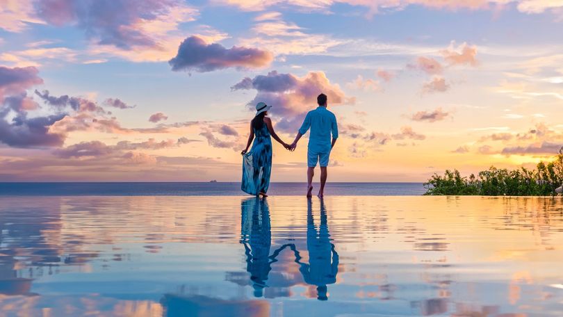 Deux personnes se tenant par la main sur une plage réfléchissante au coucher du soleil, sous des nuages pastel.