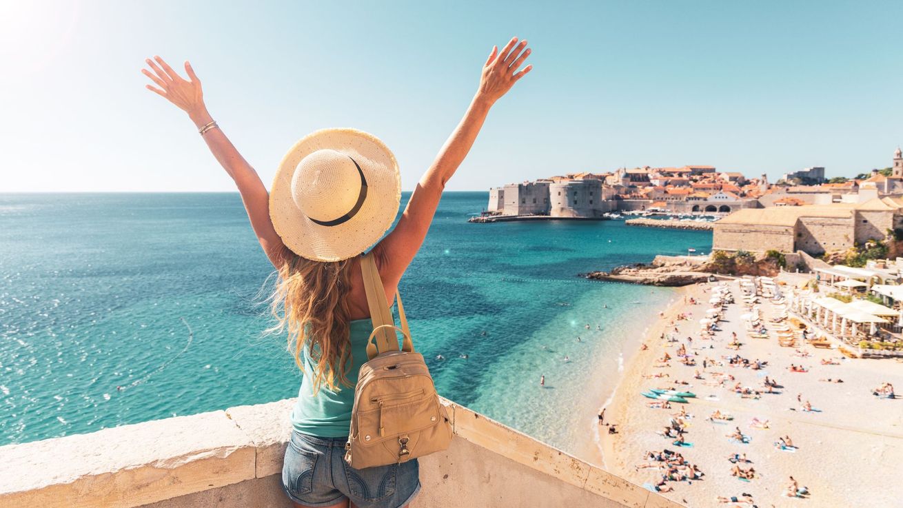 Femme au chapeau de paille, les bras levés, admirant une plage turquoise et une ville en bord de mer.