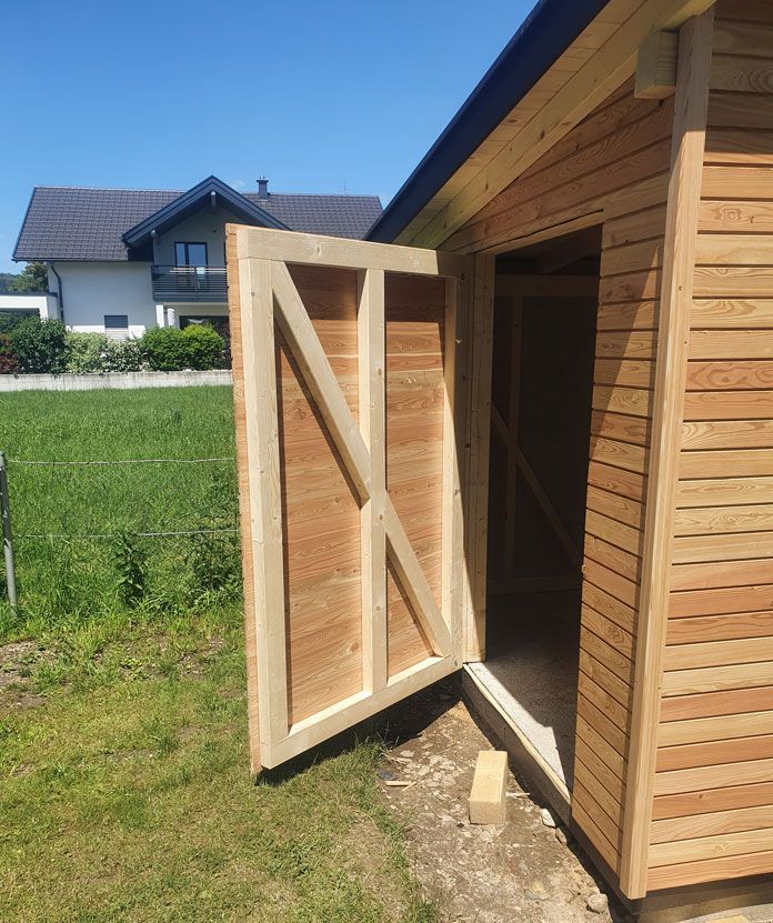 Offene Tür zum Holzschuppen; braune Holzkonstruktion mit Querverstrebung; sonnige Außenlage.