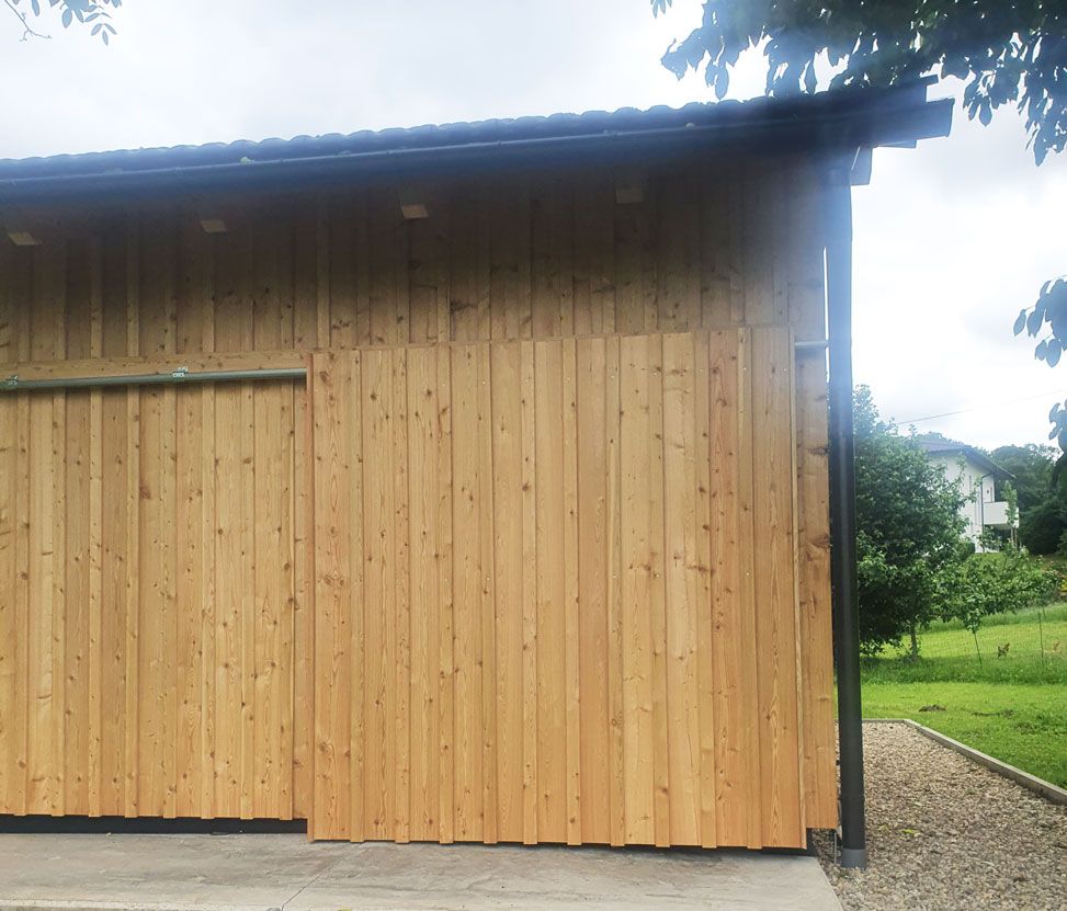 Ein Holzschuppen mit dunklem Dach und Betonfundament, der sich in einer grasbewachsenen Außenanlage befindet.