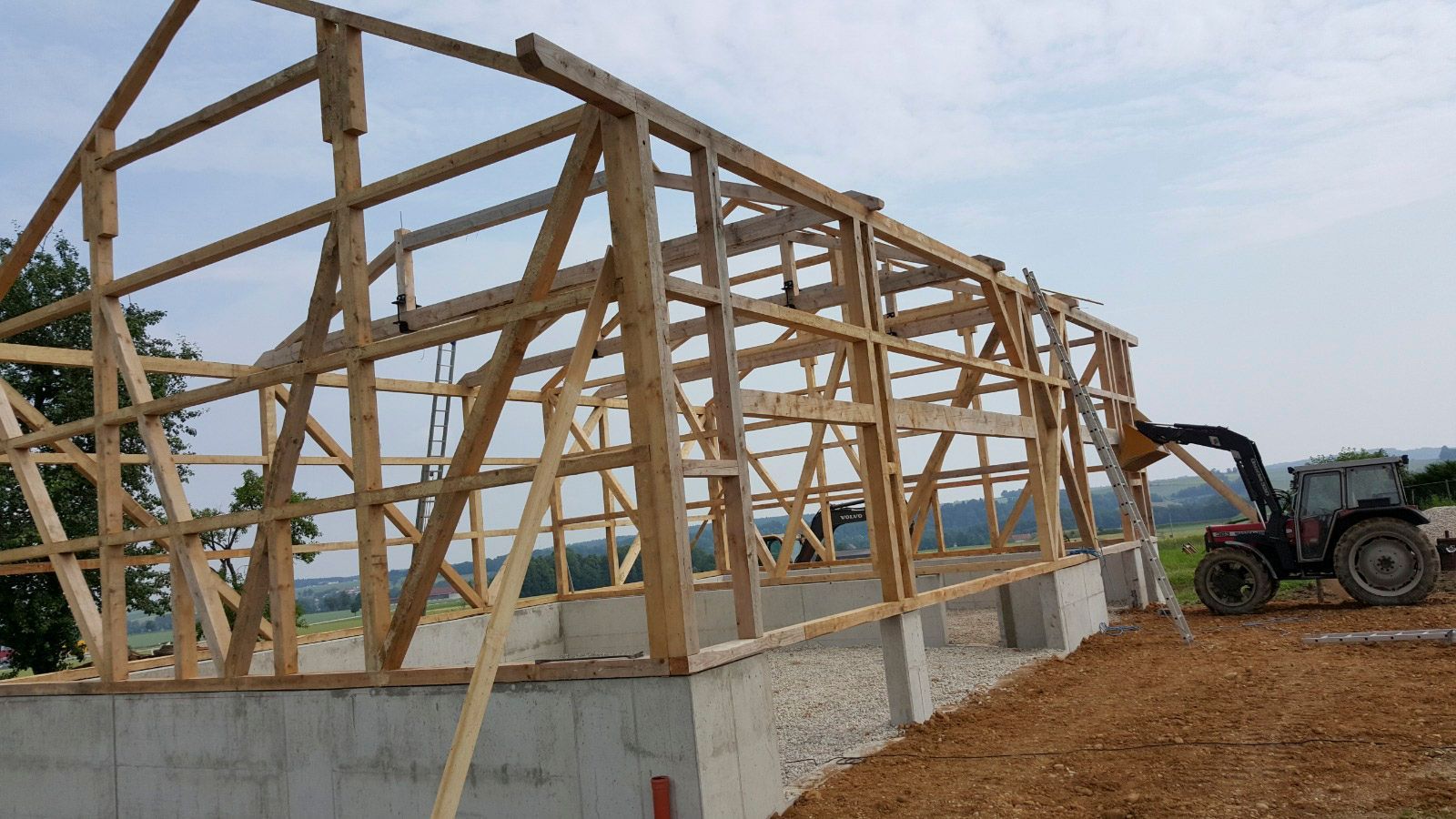 Im Bau befindliches hölzernes Scheunengerüst mit einem Traktor auf einem Betonfundament in ländlicher Umgebung.