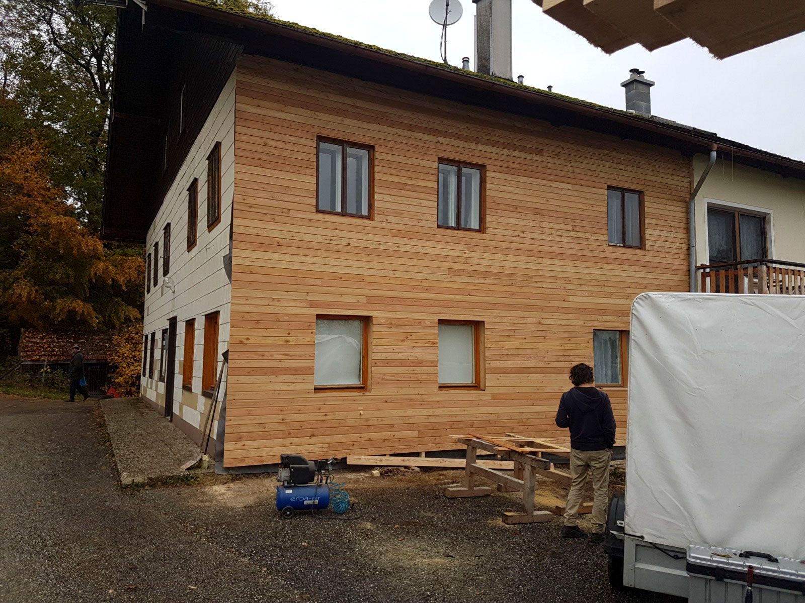 Zweistöckiges Haus mit neuer Holzverkleidung. Eine Person steht neben Werkzeugen auf der Baustelle.