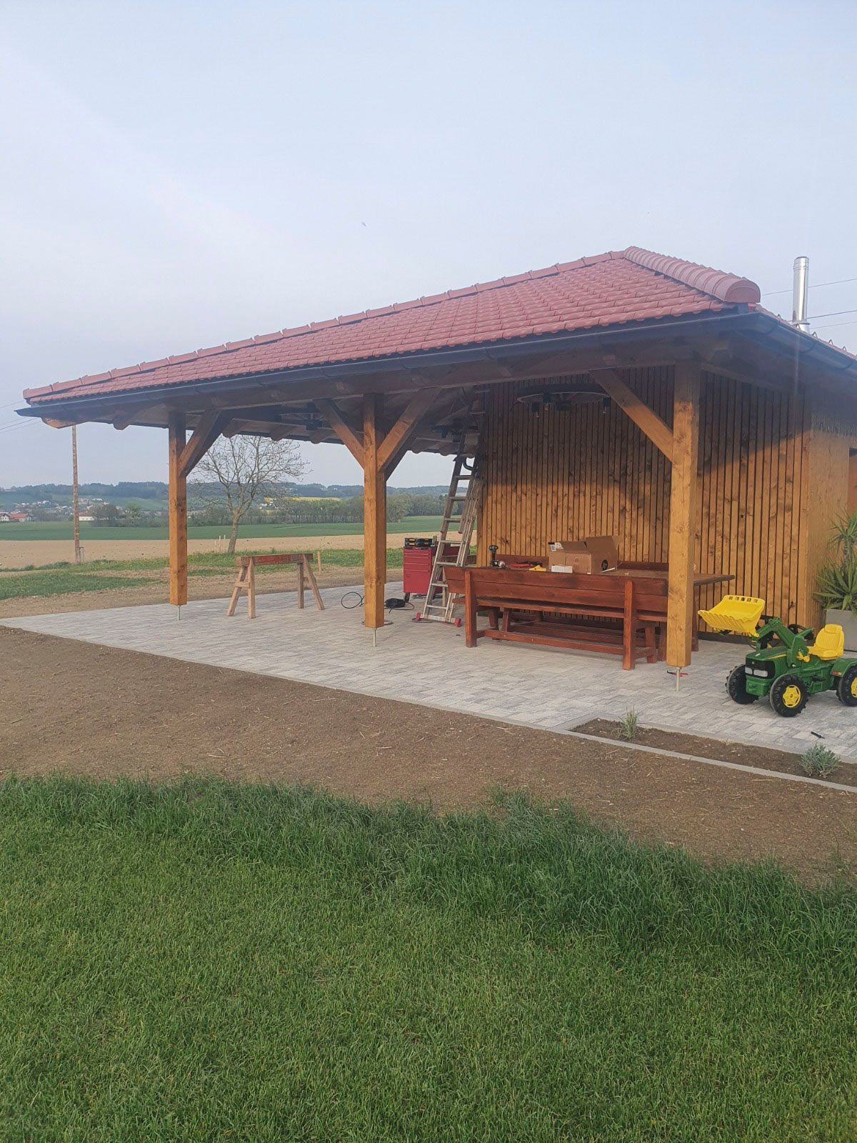 Holzpavillon mit rotem Dach, Bank und Spielzeug auf einer Steinterrasse mit Blick auf ein Feld.