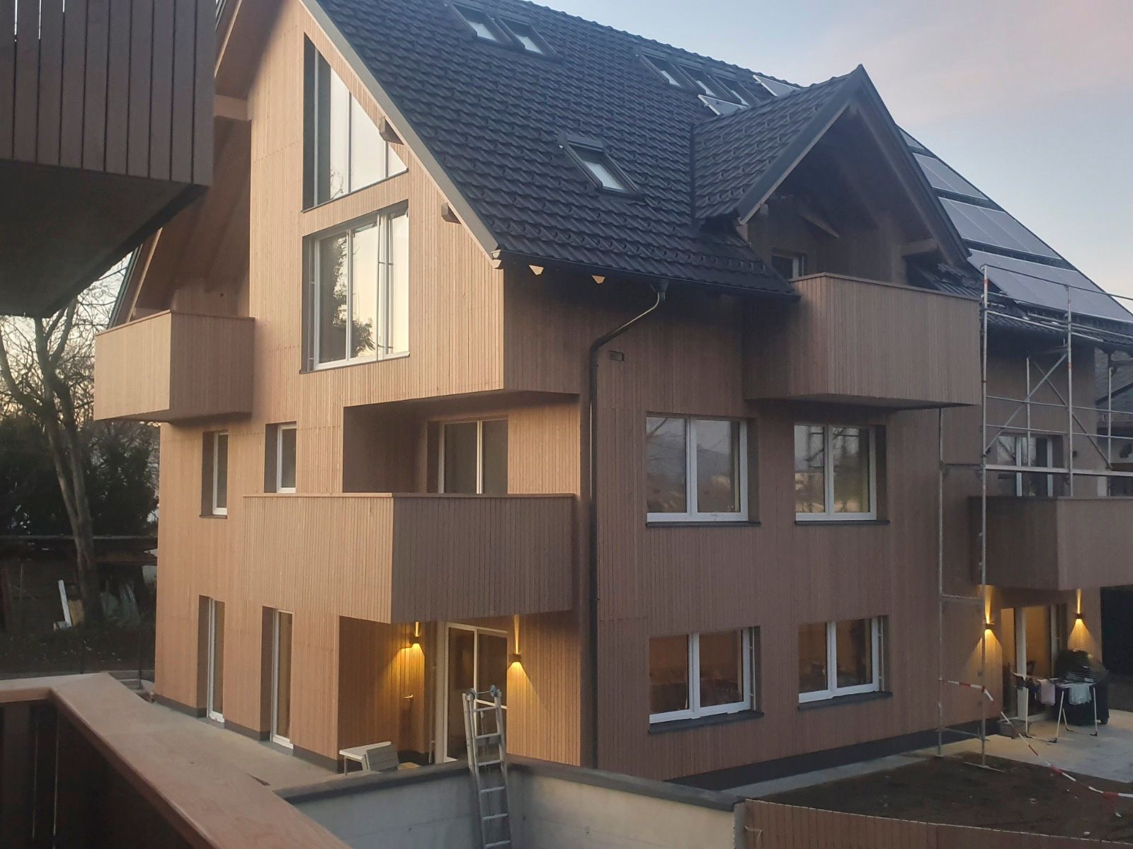 Modernes, mehrstöckiges Holzgebäude mit Balkonen, dunklem Dach und Solarpaneelen.