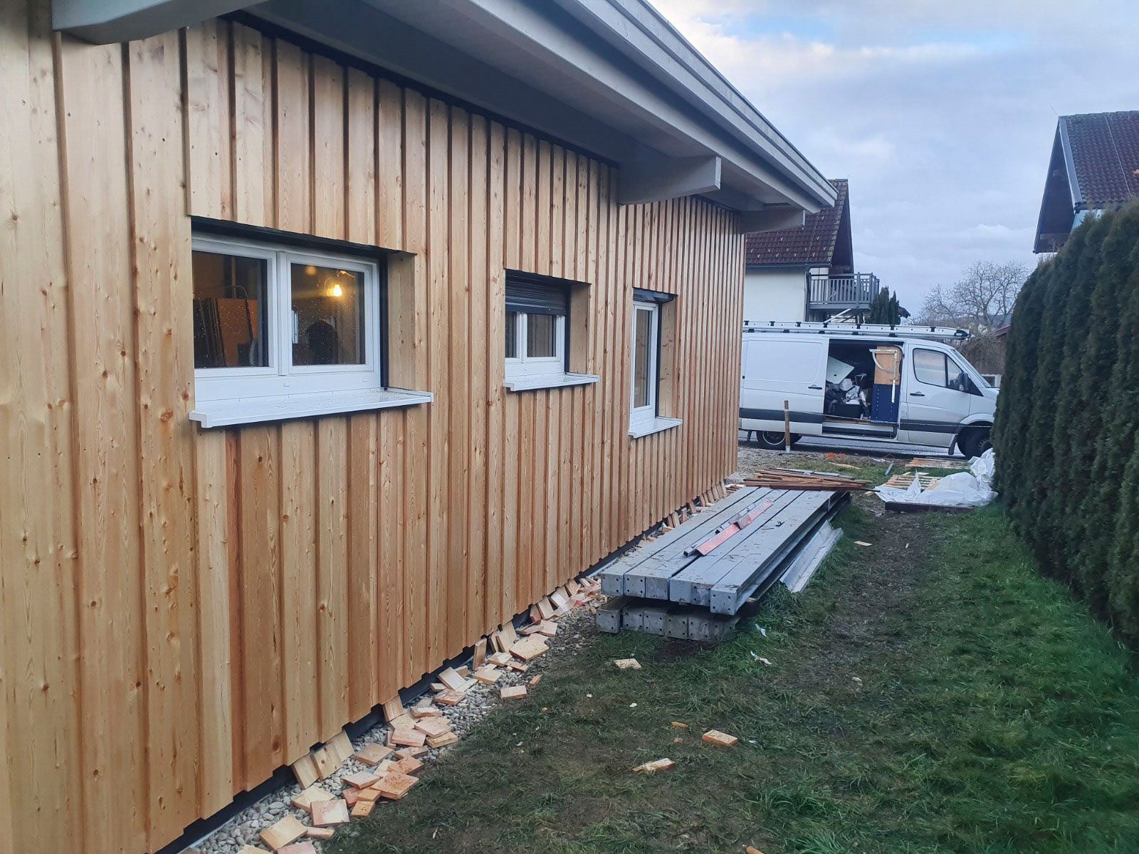 Ein mit Holz verkleidetes Gebäude mit weiß gerahmten Fenstern. Baumaterialien und ein Lieferwagen 