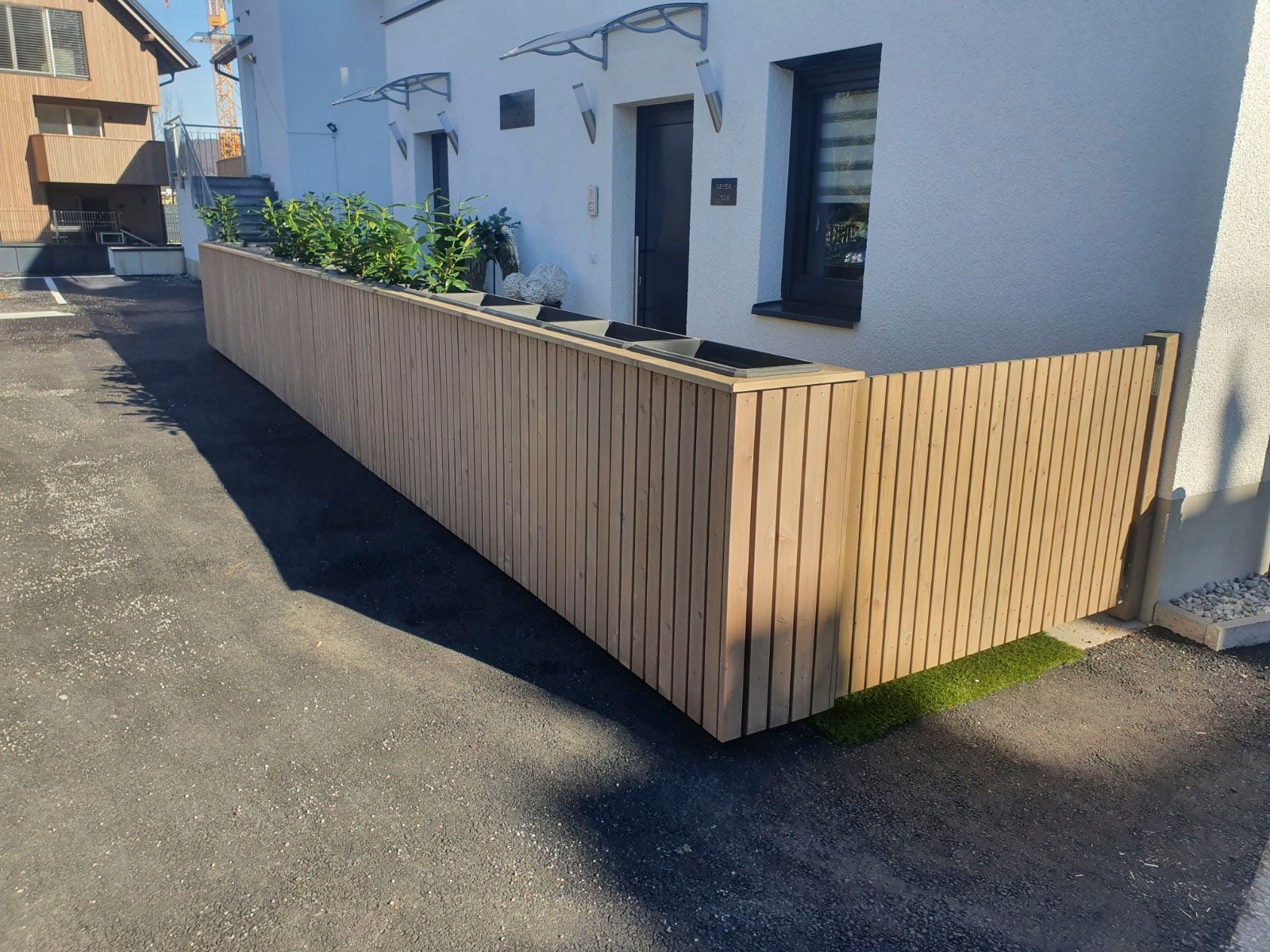 Ein hölzerner Lattenzaun mit Pflanzen vor einem weißen Gebäude mit schwarzer Tür und schwarzen Fenstern.