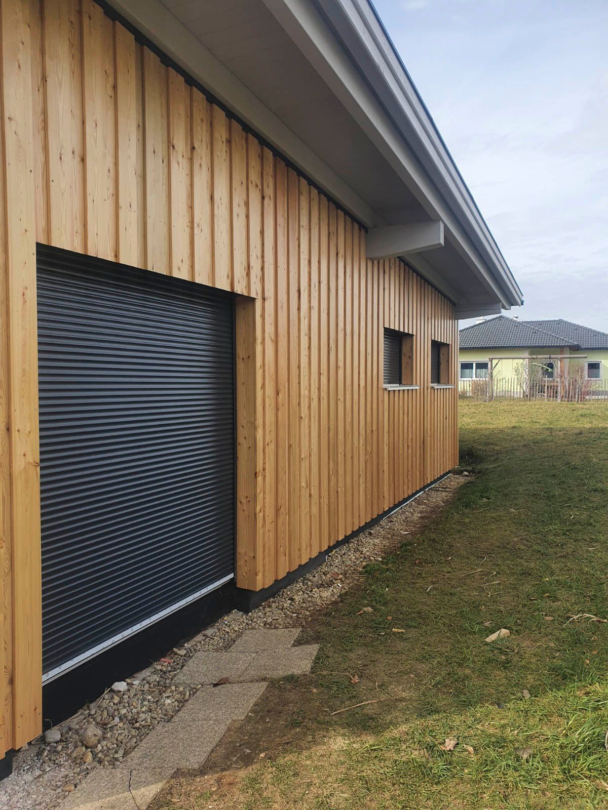 Außenansicht eines Gebäudes mit Holzverkleidung, einem geschlossenen schwarzen Rollladen und einem kleinen Fenster.
