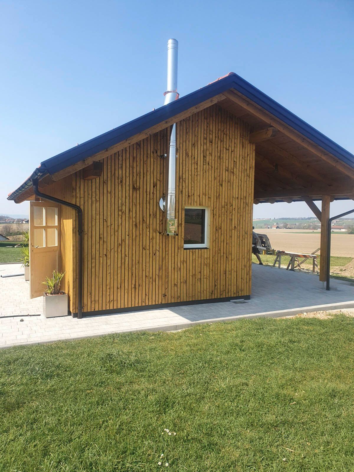 Eine Holzhütte mit Kamin auf einer Rasenfläche unter blauem Himmel, mit überdachter Terrasse.