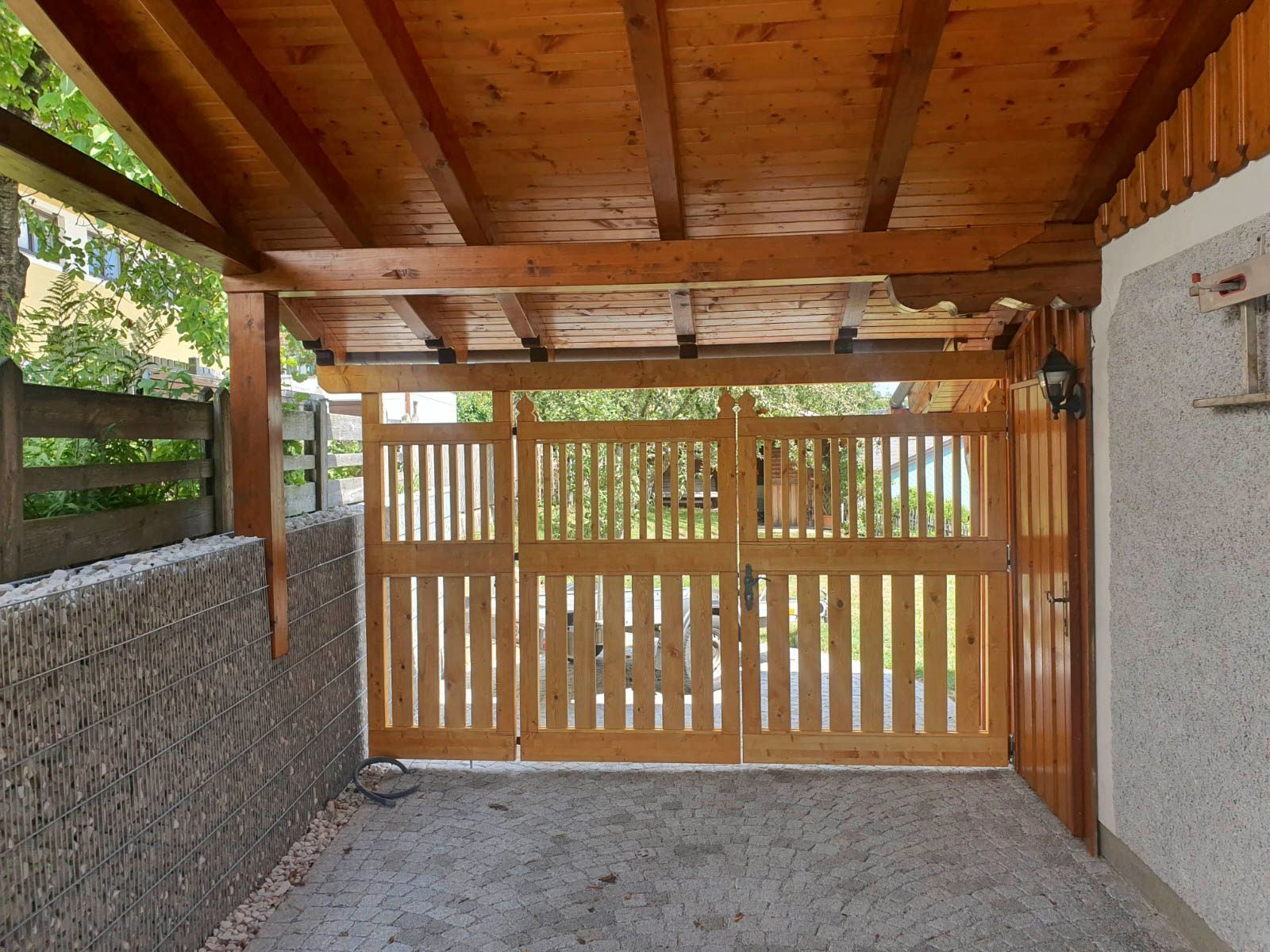 Carport aus Holz mit Tor. Hellbraunes Holz, Kiesboden.