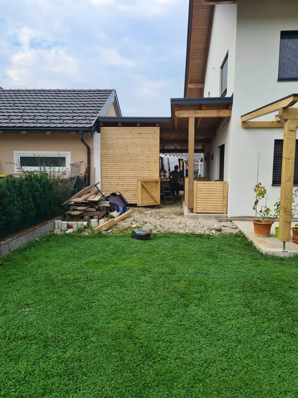 Hinterhof mit Bebauung: Holzpergola, Haus, kleines Gebäude und grüner Rasen.