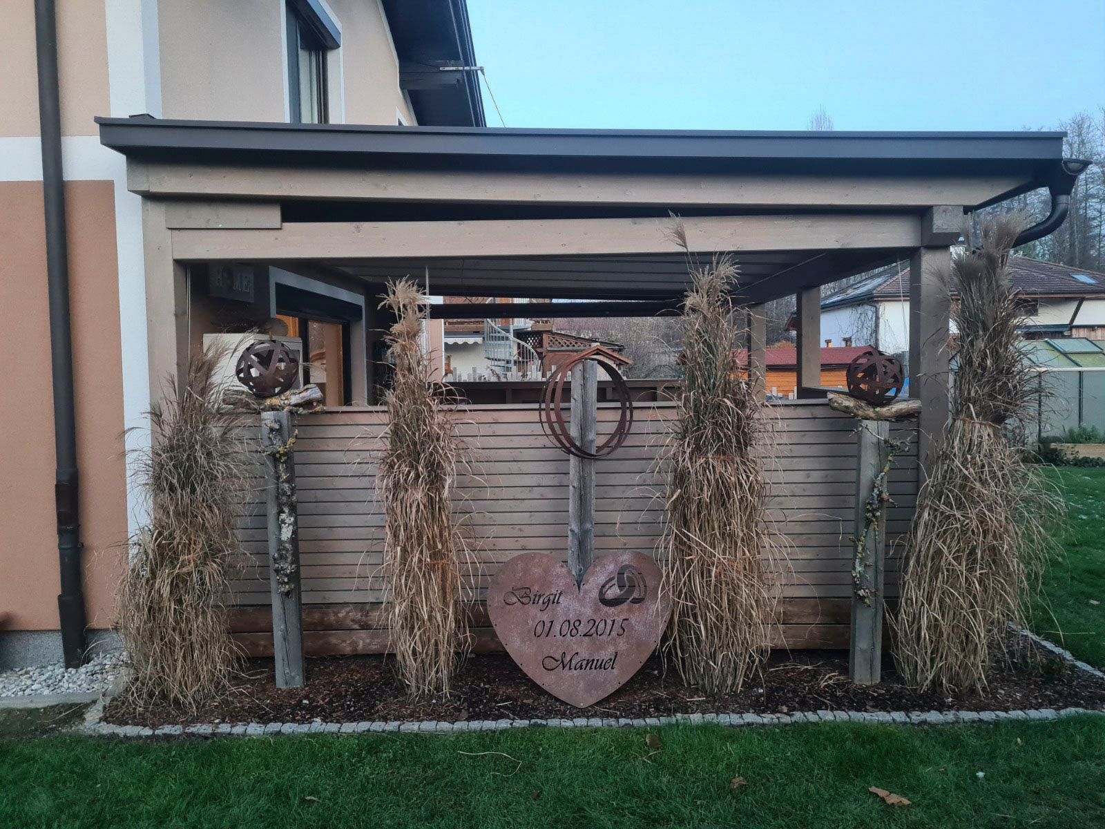 Eine mit getrockneten Gräsern, Metallherzen und einem Schild verzierte Holzpergola steht auf einem Rasen.